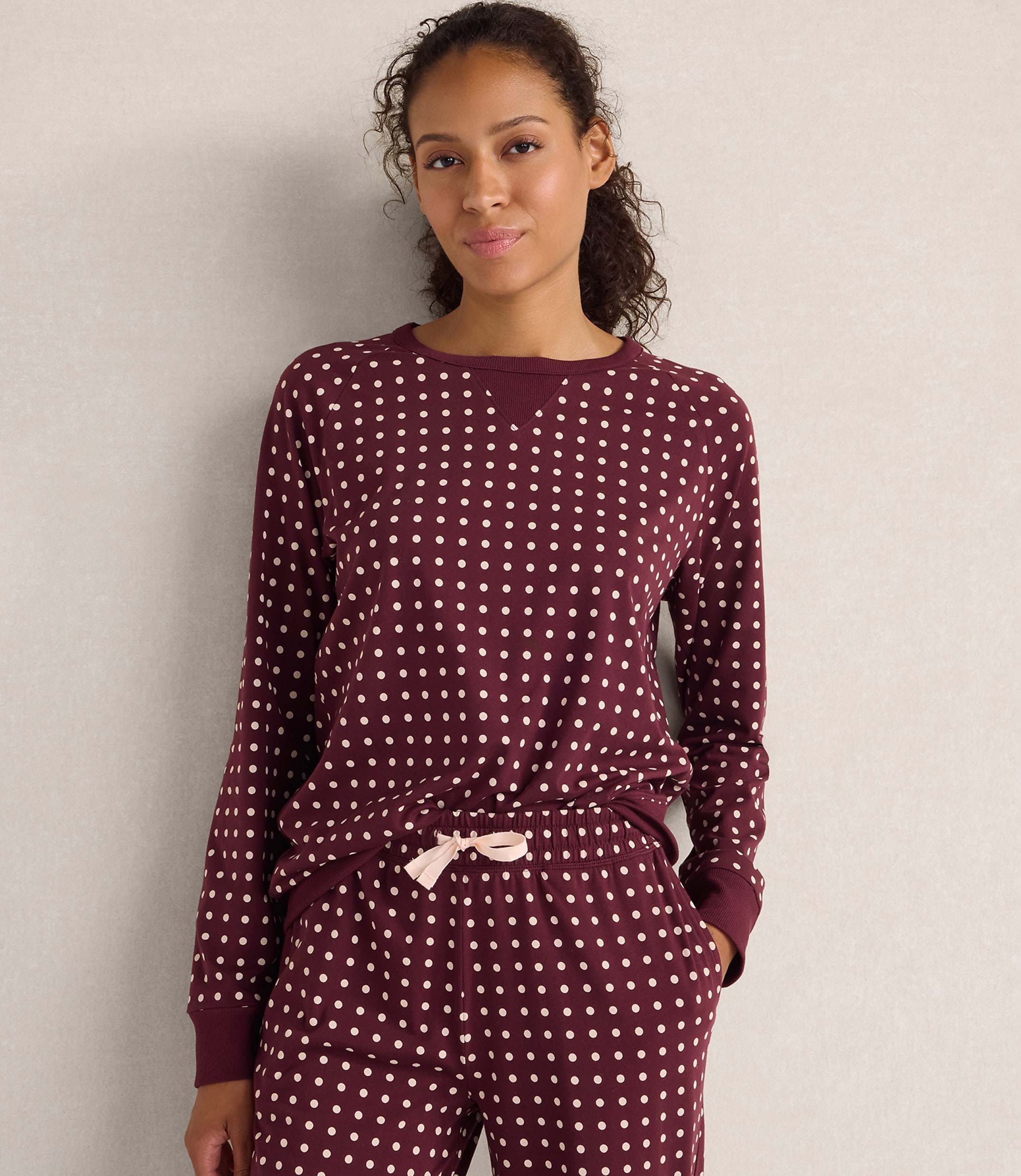 A woman wearing maroon polka-dot pajamas stands against a plain, light-colored wall.