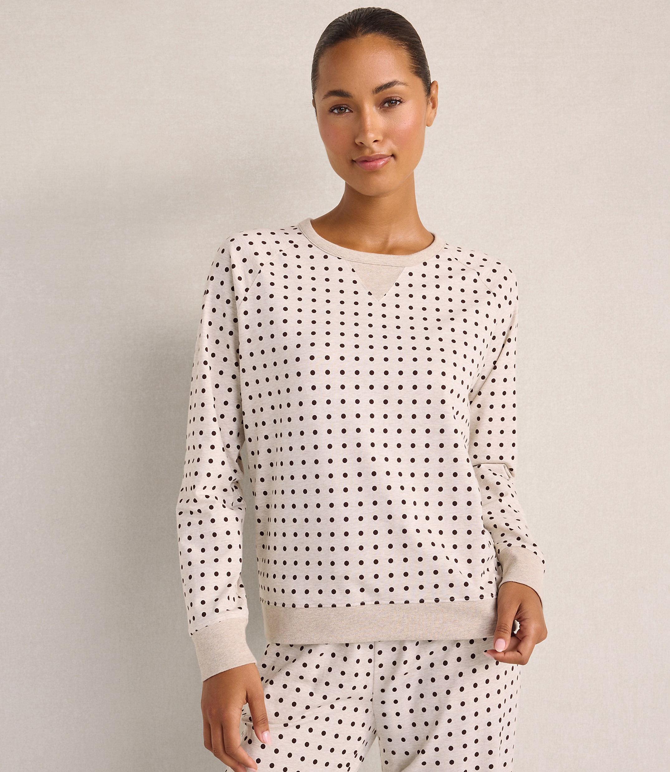 A woman wearing a cream-colored polka-dot pajama set stands against a plain, neutral-toned wall.
