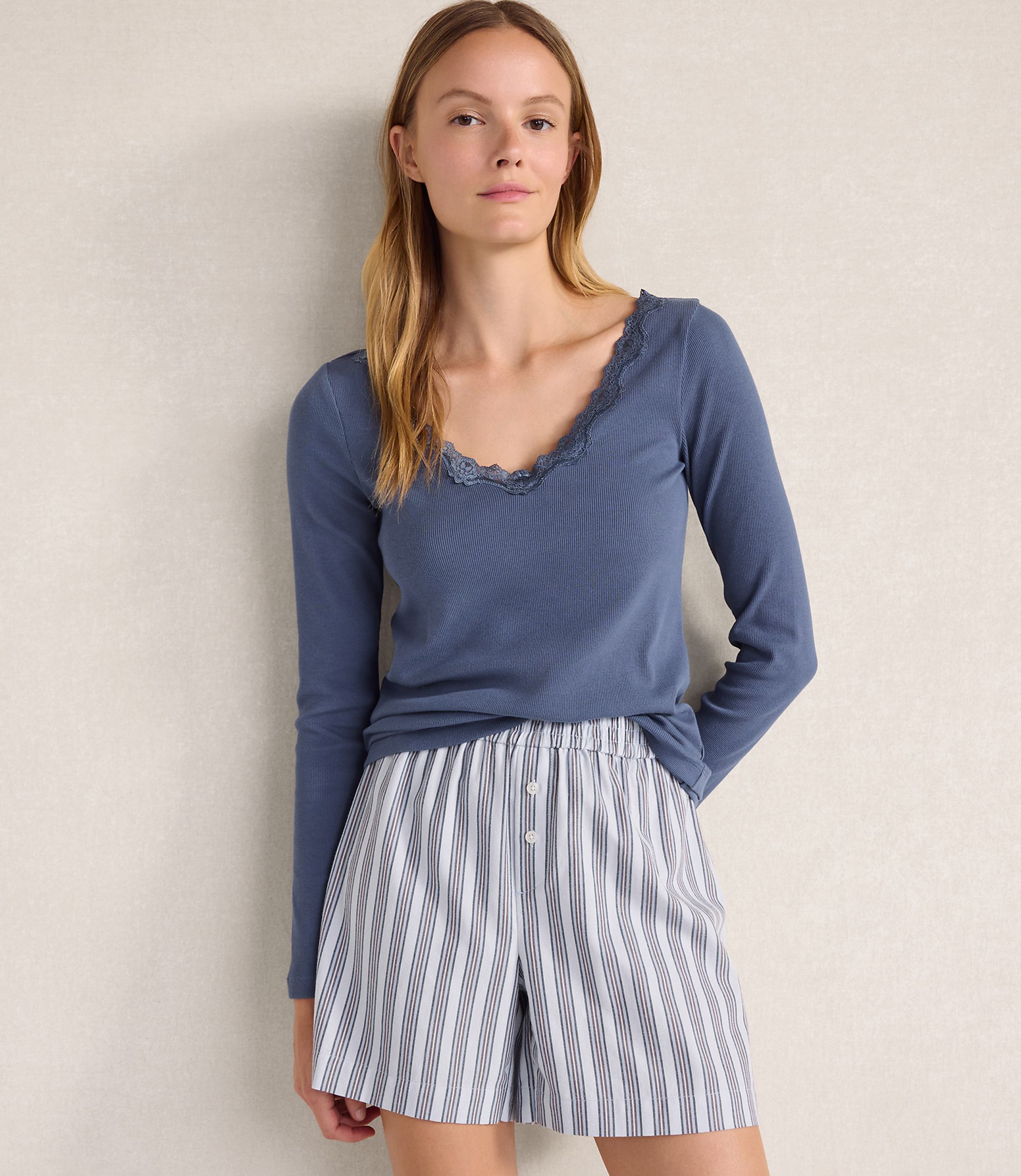 A woman wearing a blue long-sleeved top and striped shorts stands against a plain, light-colored wall.