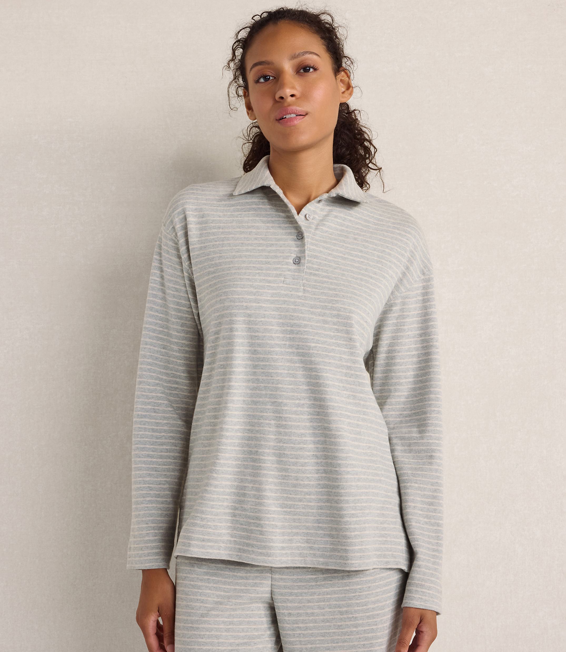 A woman wearing a grey striped long-sleeved polo shirt and matching pants stands against a plain, neutral-colored wall.