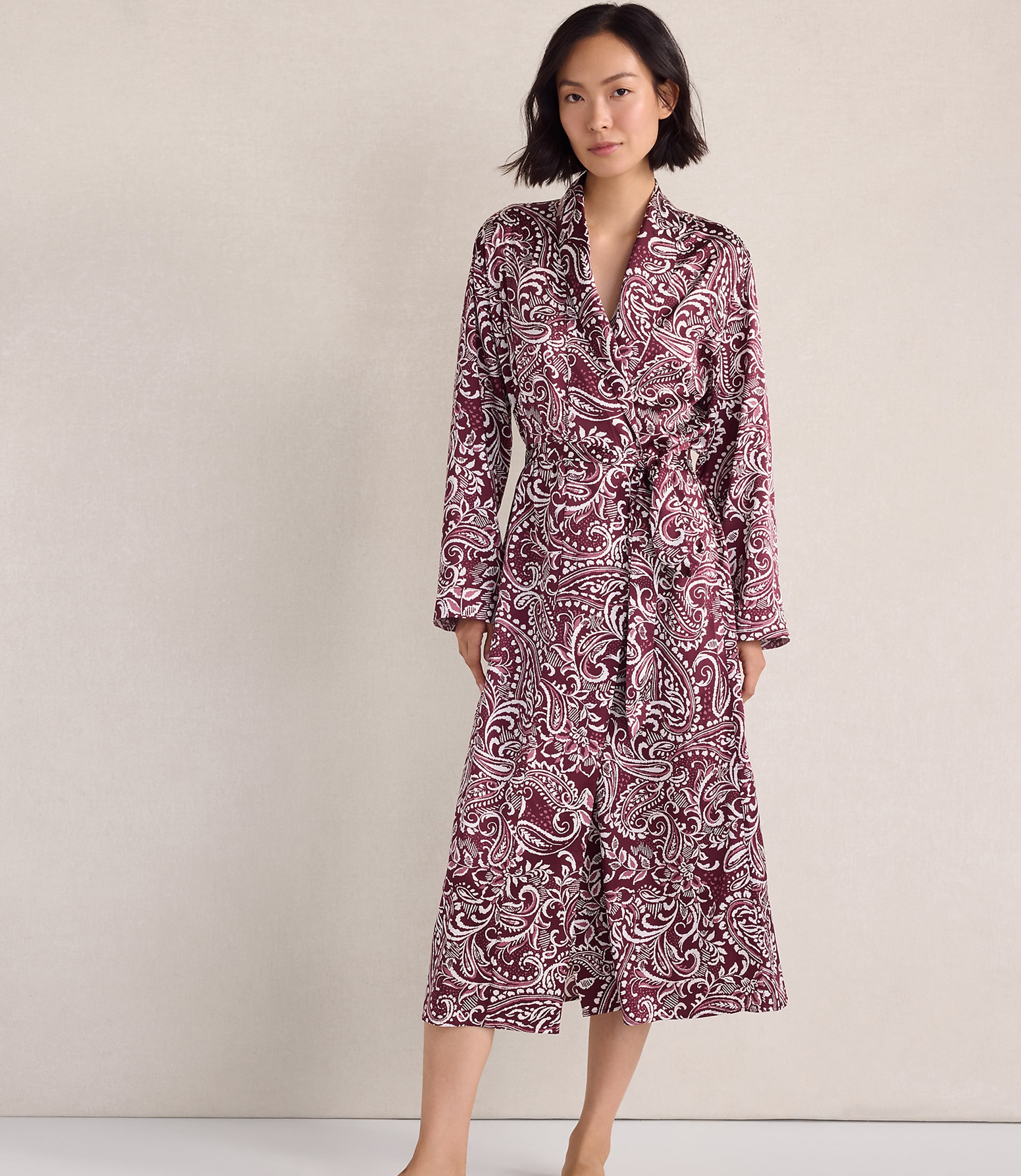 A woman wearing a maroon and white paisley print robe stands against a plain, neutral-colored wall.