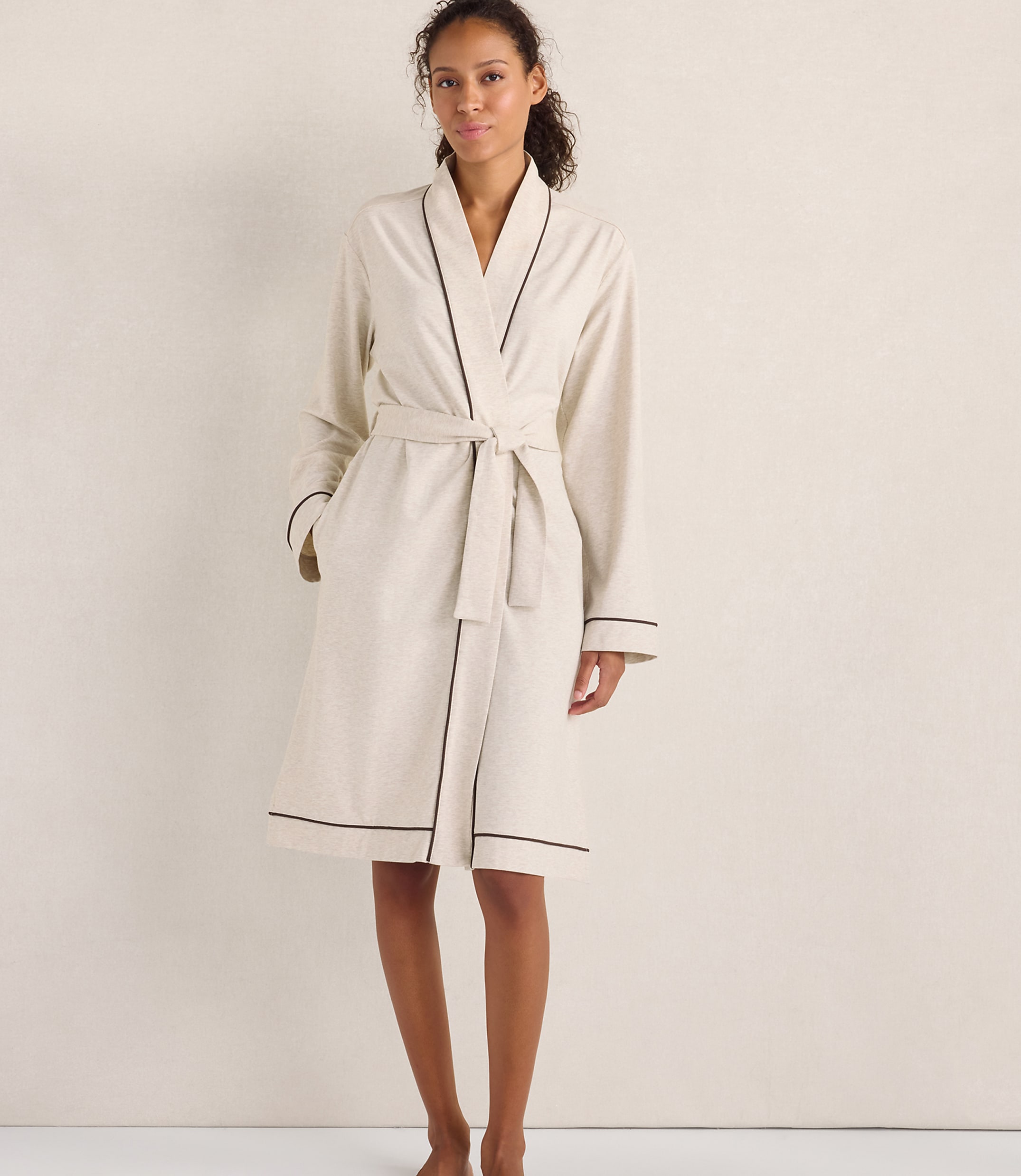 A woman wearing a cream-colored robe with dark piping stands against a plain, neutral-toned wall.