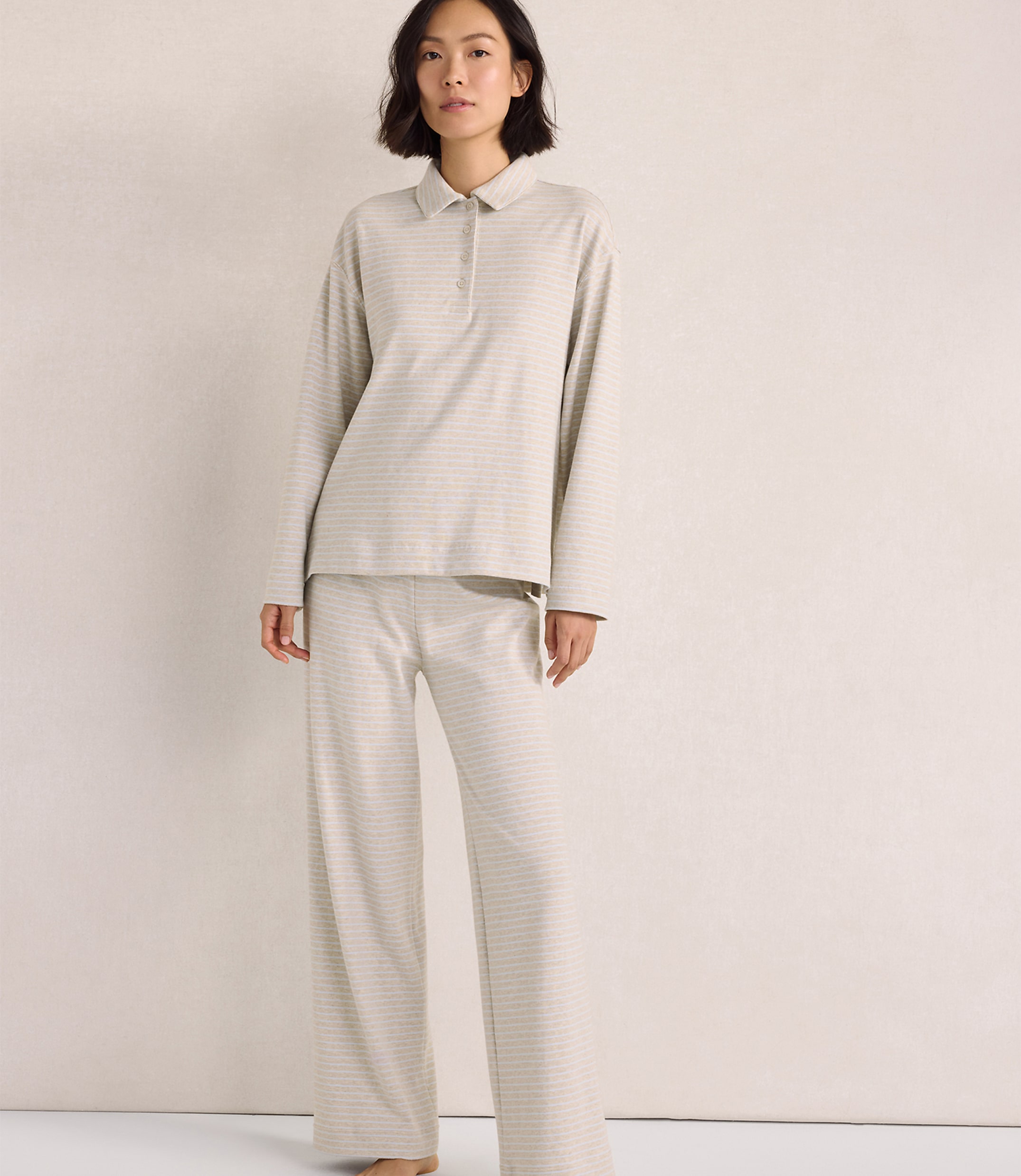 A woman wearing a light-colored, striped long-sleeved polo shirt and matching wide-leg pants stands against a plain, neutral-toned wall.