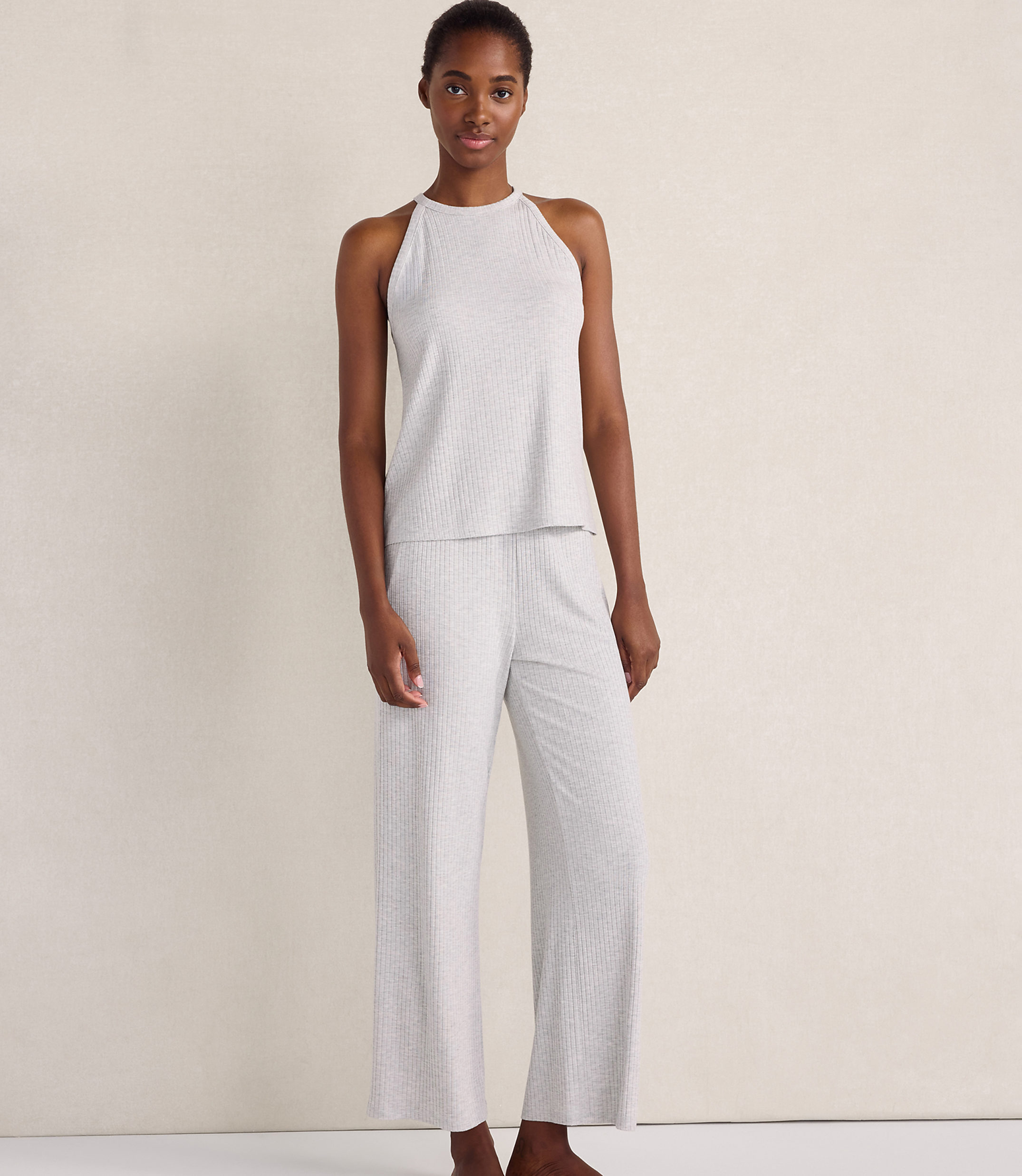 A woman wearing a light grey ribbed sleeveless top and matching wide-leg pants stands against a plain, neutral-toned wall.