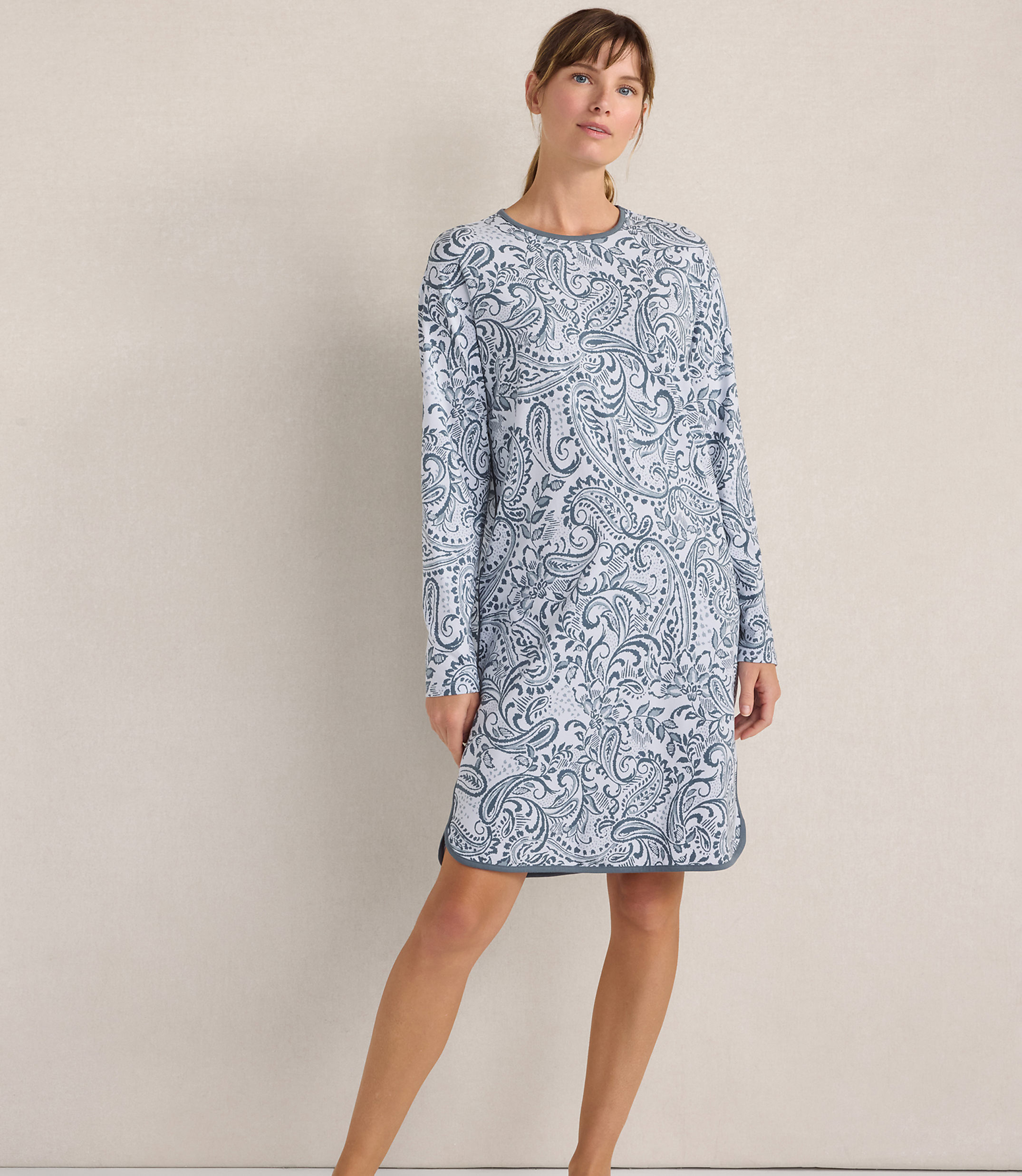 A woman wearing a blue and white paisley print long-sleeved dress stands against a plain, neutral-colored wall.