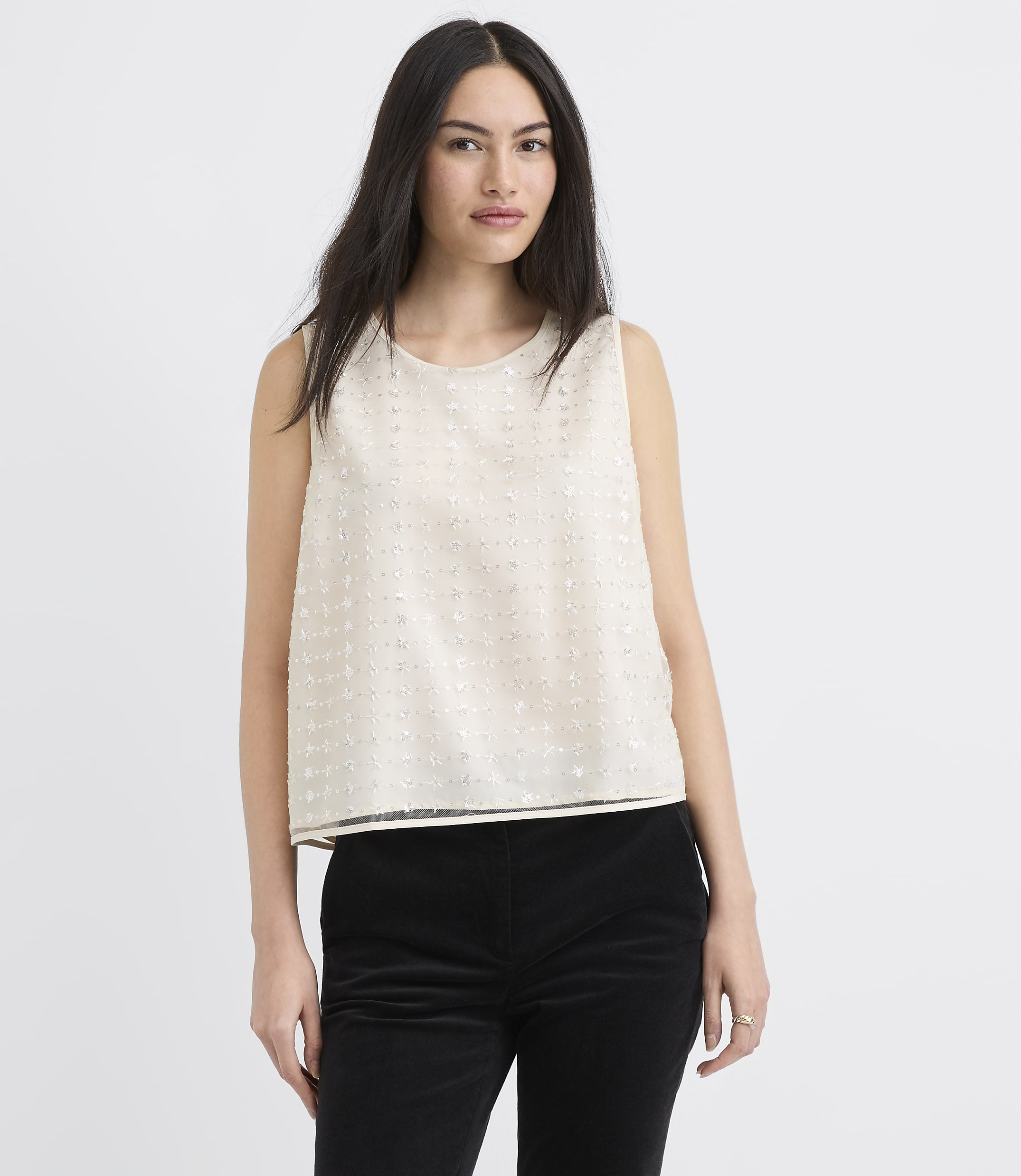 A woman wearing a sleeveless white textured top and black trousers stands against a plain white background.