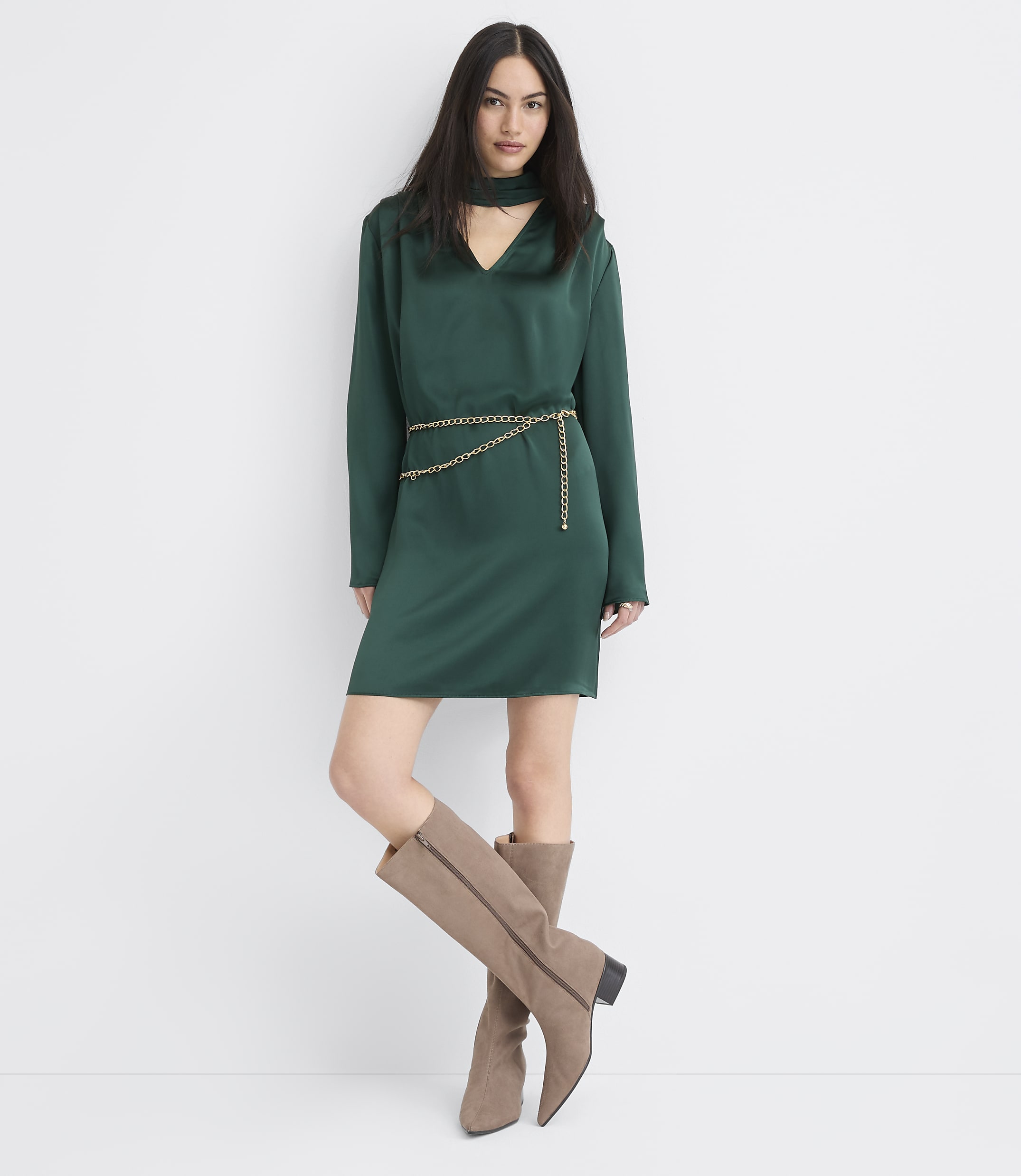A woman wearing a green satin dress with a gold chain belt and taupe boots stands against a plain white background.