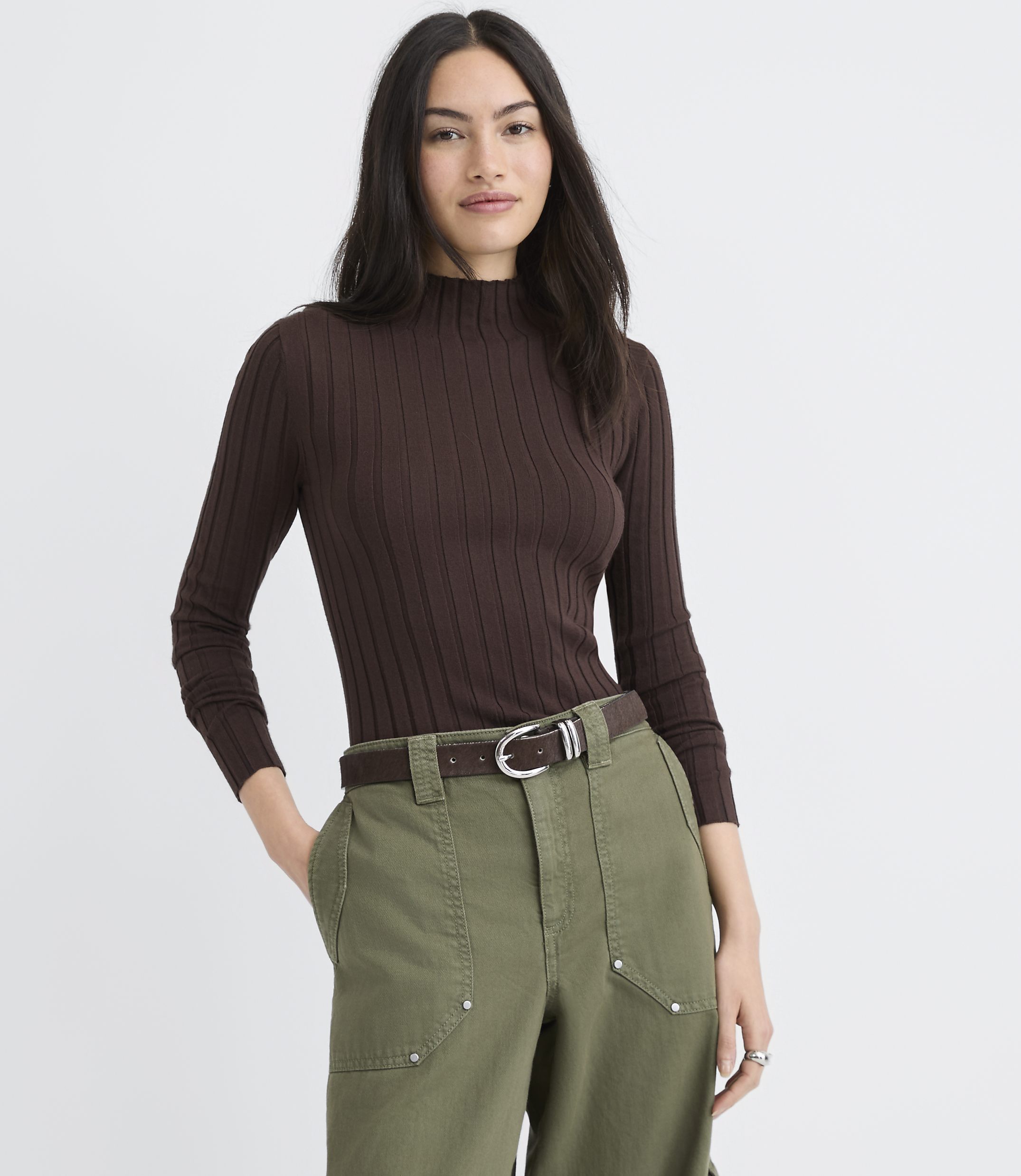 A woman wearing a brown ribbed turtleneck and olive green pants stands against a plain white background.