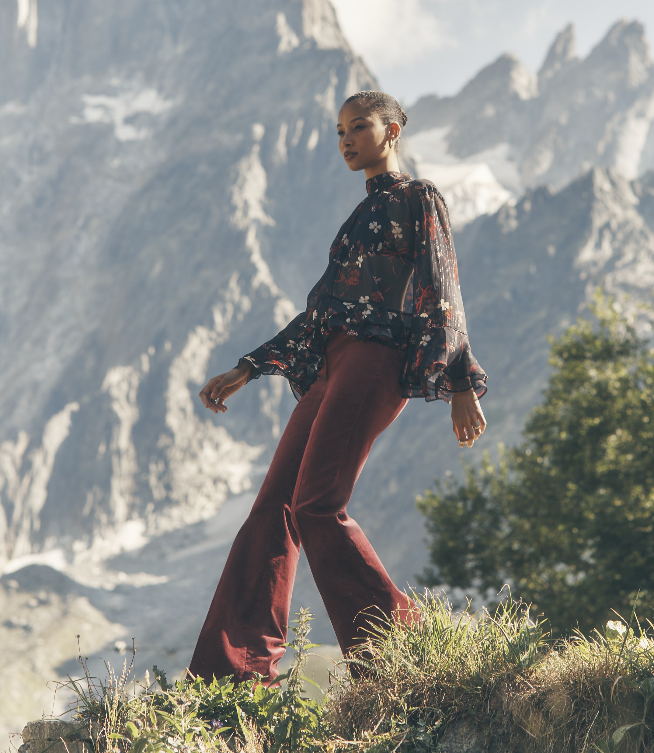 A woman in a floral blouse and red trousers stands on a grassy ledge against a backdrop of towering, jagged mountains.