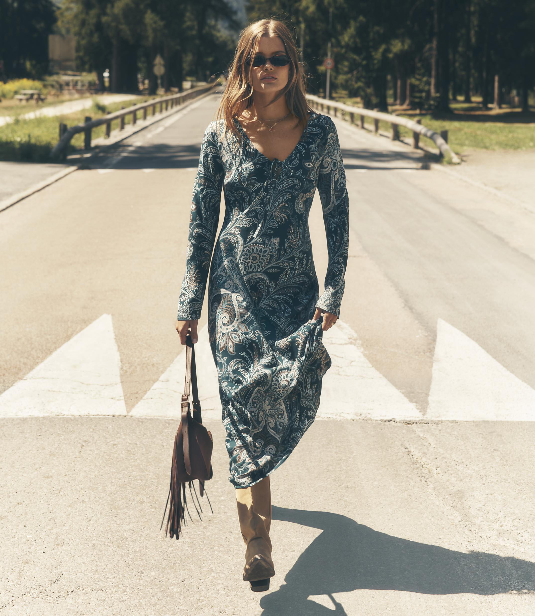 A woman wearing a long, patterned blue dress and sunglasses walks down a sunny road lined with trees and wooden railings.
