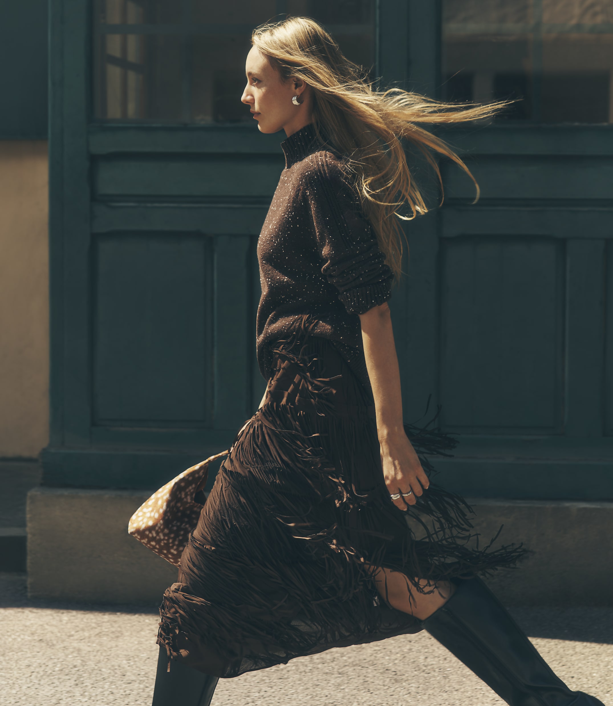 A blonde woman in a brown knit top and fringed skirt walks past a dark green paneled wall.