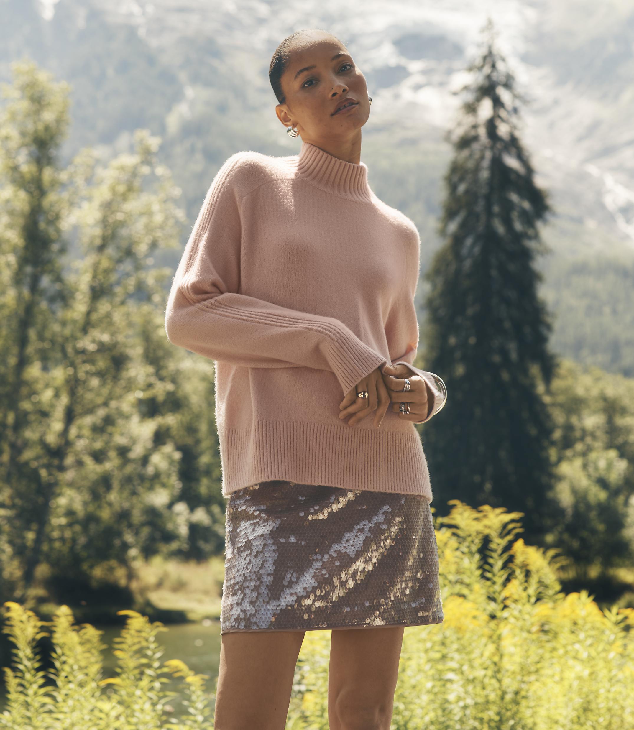 A woman wearing a pink sweater and a shimmering skirt stands in a lush green field with mountains and evergreen trees in the background.