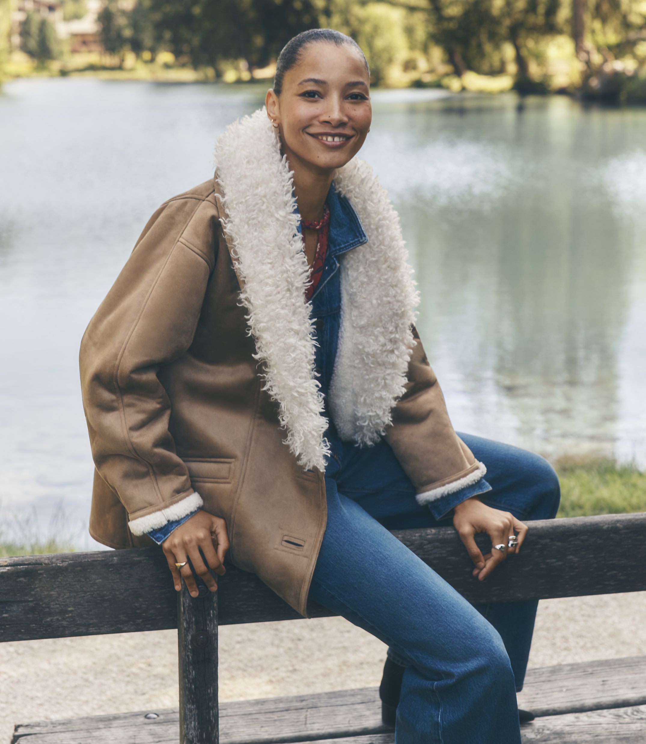 A smiling woman wearing a tan shearling coat and blue jeans sits on a wooden bench in front of a calm lake and lush greenery.
