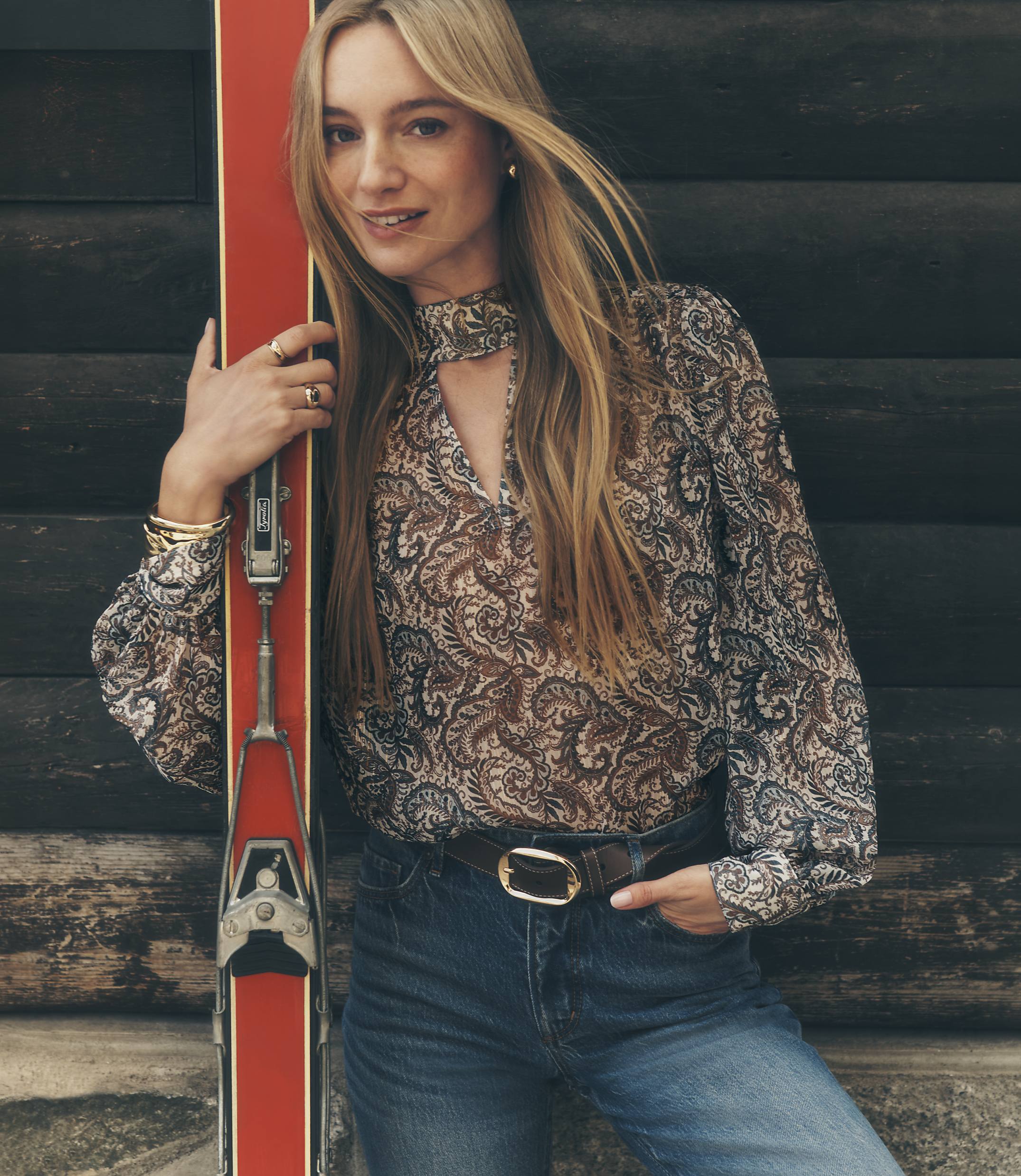 A blonde woman in a paisley blouse and jeans leans against a red ski in front of a dark wooden log wall.