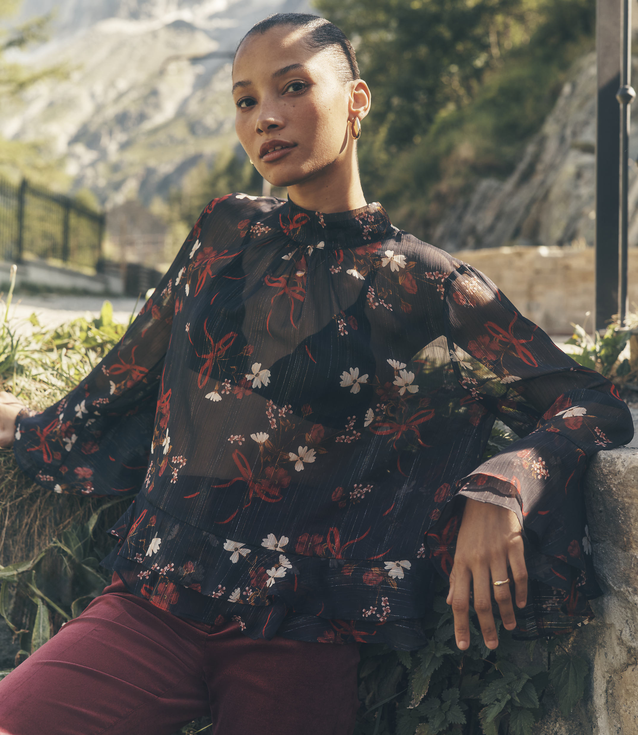 A woman wearing a sheer floral blouse and maroon trousers poses against a backdrop of rocky mountains and greenery.
