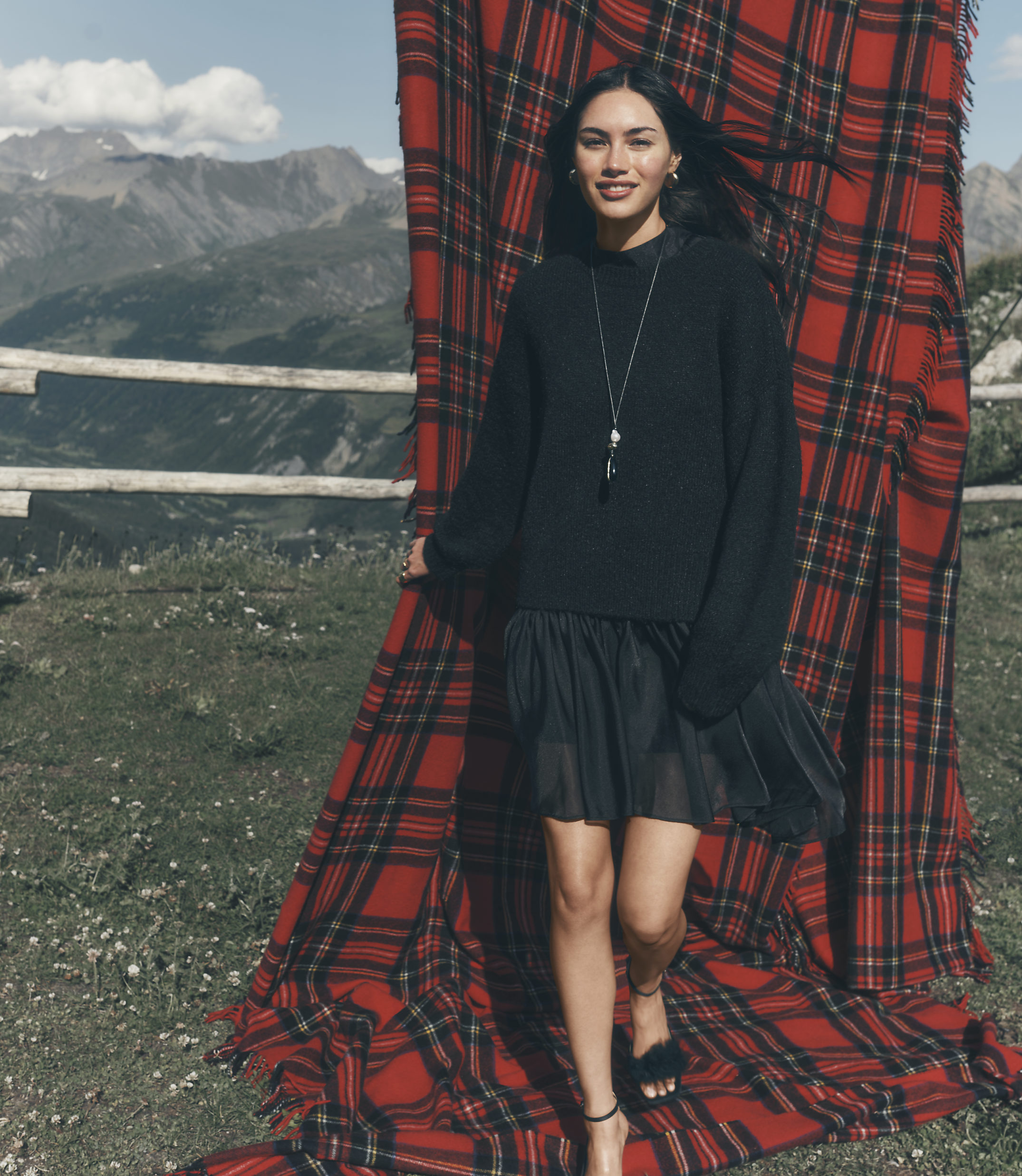 A woman in a black sweater and skirt stands before a large red plaid blanket with a mountainous landscape in the background.