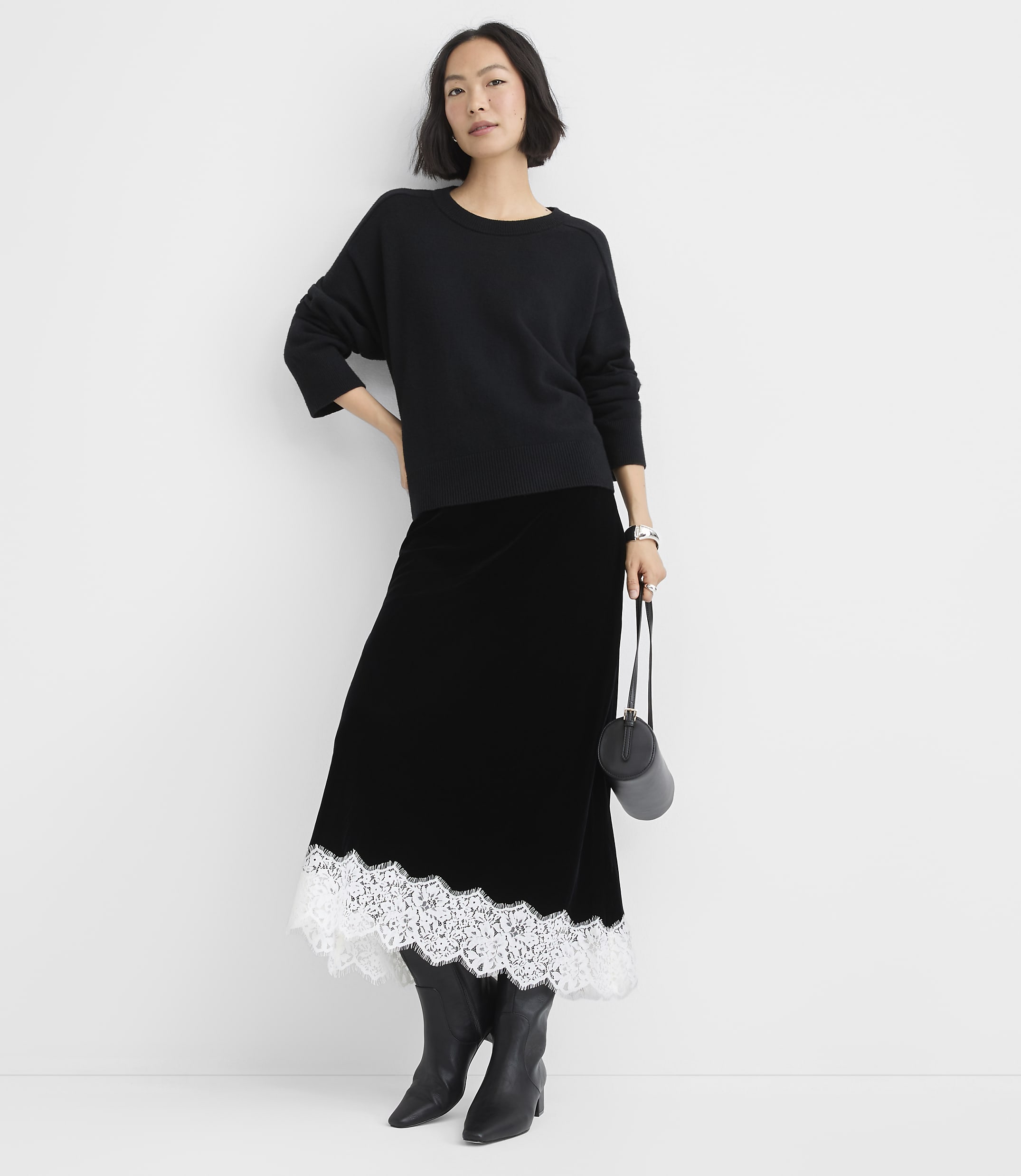 A woman wearing a black sweater, a long black skirt with white lace trim, and black boots stands against a plain white background.