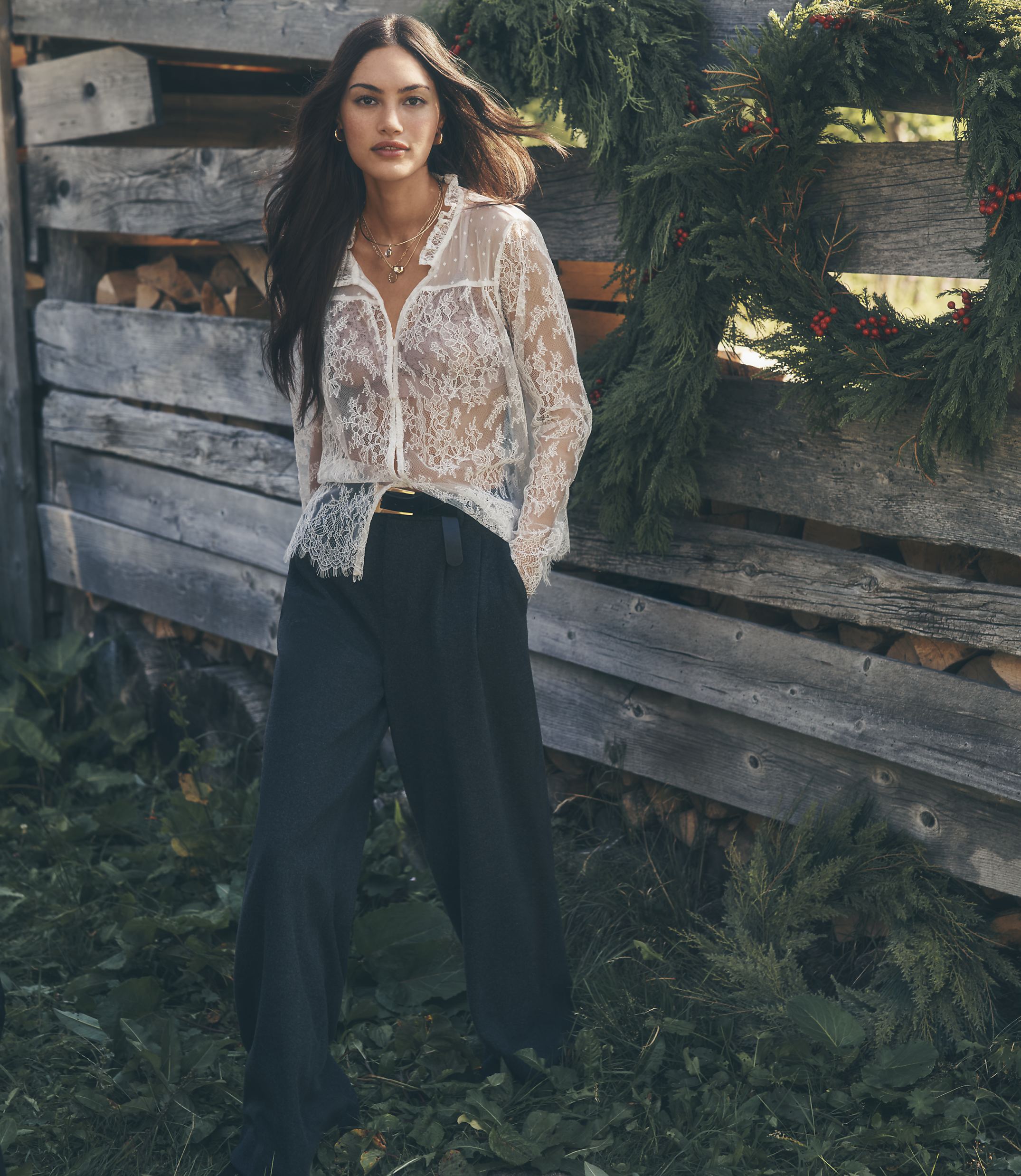 A woman in a white lace blouse and black trousers stands in the foreground against a background of a rustic wooden fence adorned with evergreen branches.