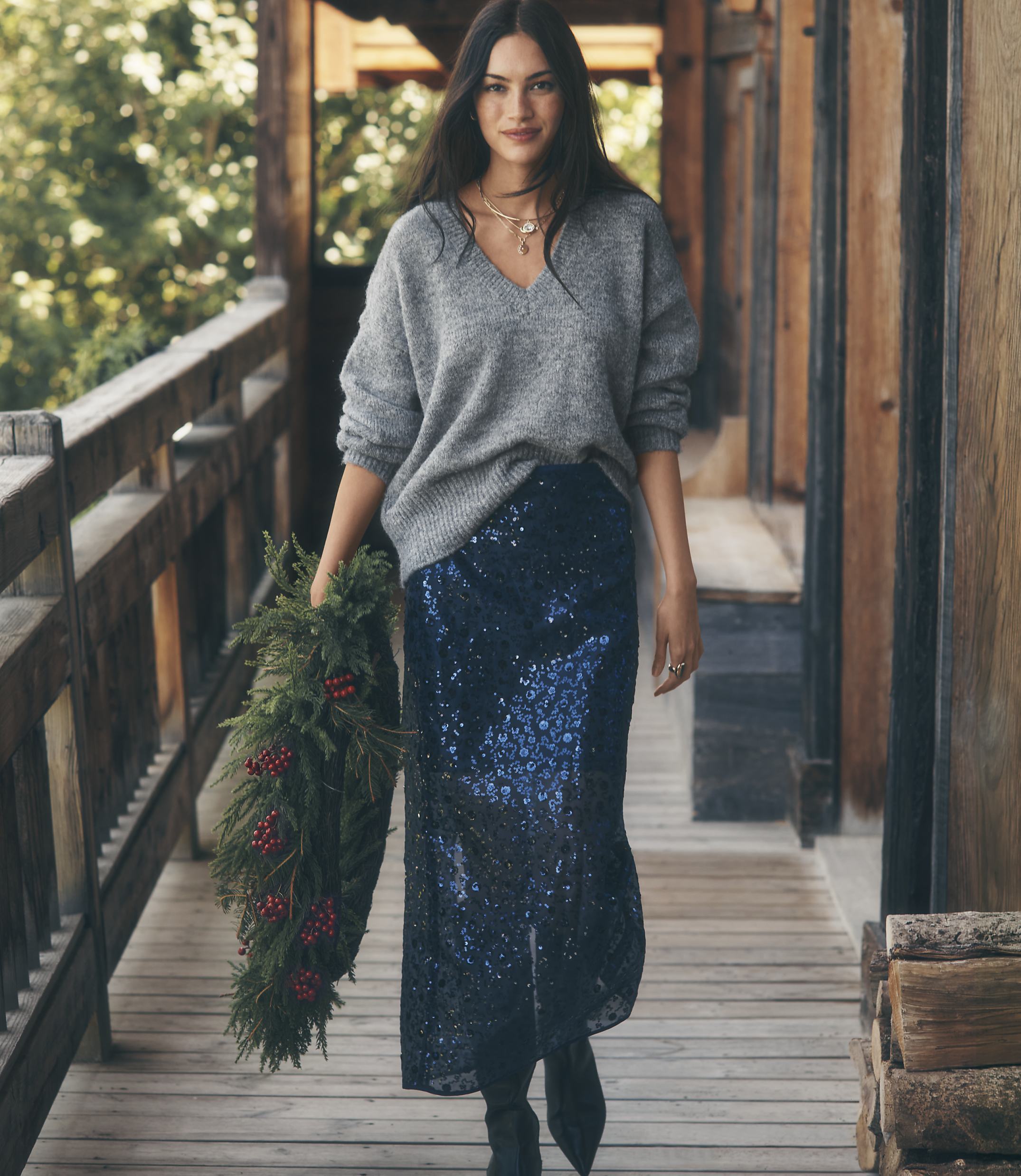 A woman wearing a grey sweater and a navy sequined skirt holds a holiday wreath while walking along a rustic wooden porch.