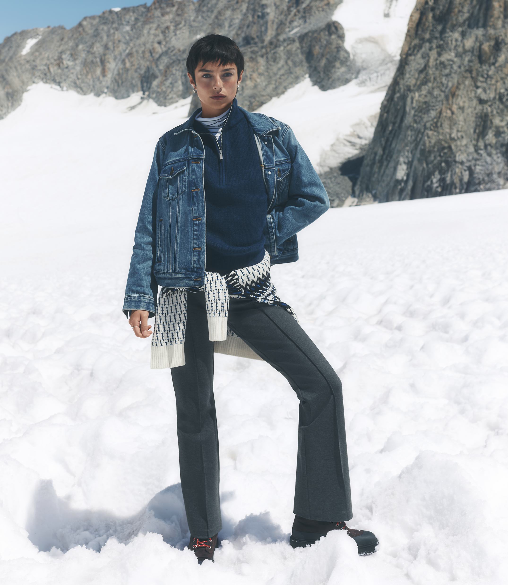 A woman wearing a denim jacket and dark trousers stands in a snowy foreground against a backdrop of rugged, snow-capped mountains.