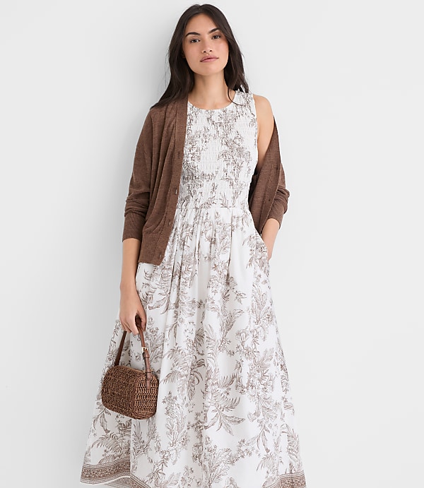 A young woman wearing a white floral dress and a brown cardigan stands in front of a plain white background, holding a woven handbag.
