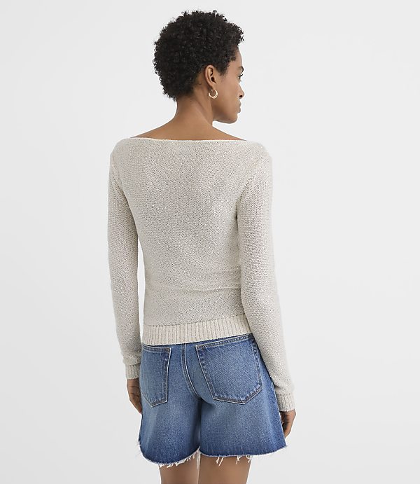 A woman in a light-colored off-the-shoulder sweater and denim shorts stands against a plain white background.