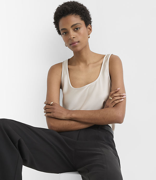 A woman in a beige tank top and black pants sits with her arms crossed against a plain white background.