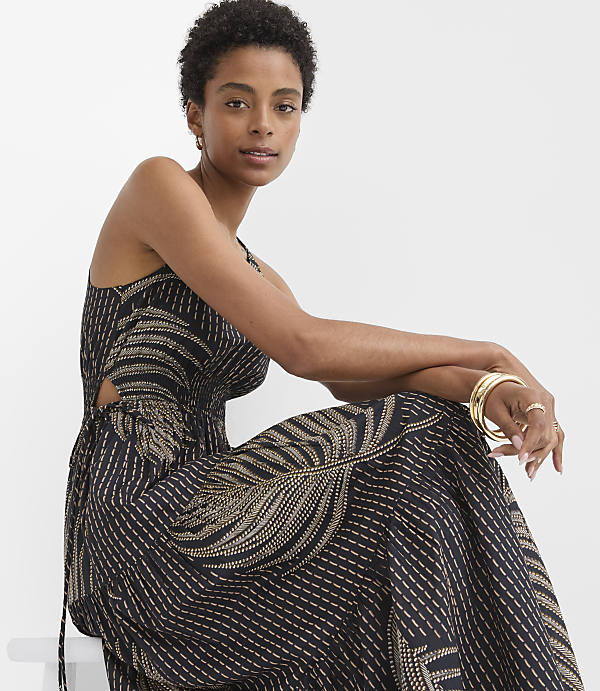 A woman in a black and gold patterned maxi dress with cutouts sits against a plain white background.