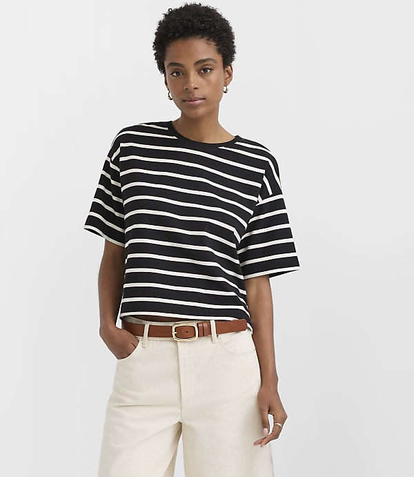 A woman in a black and white striped t-shirt and cream pants stands against a plain white background.