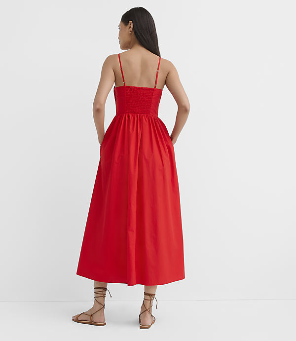 A woman in a red smocked-bodice midi dress stands with her hands in her pockets against a plain white background.
