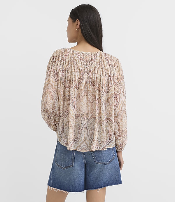 A woman in a patterned blouse and denim shorts stands against a plain, light-colored background.