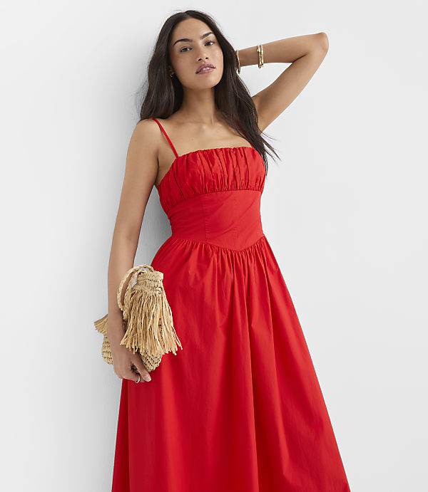 A woman in a red dress holds a straw clutch in front of a plain white background.