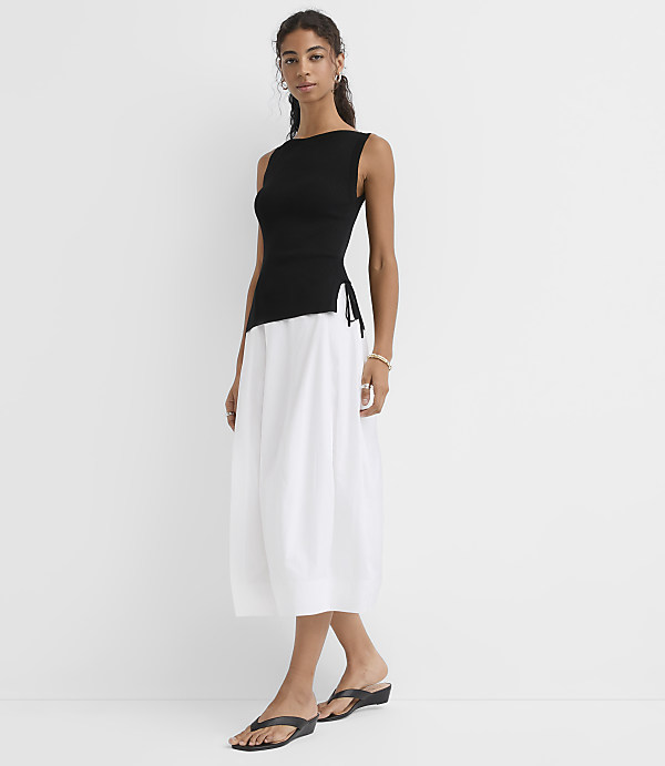 A woman in a black tank top and white skirt stands against a plain white background.