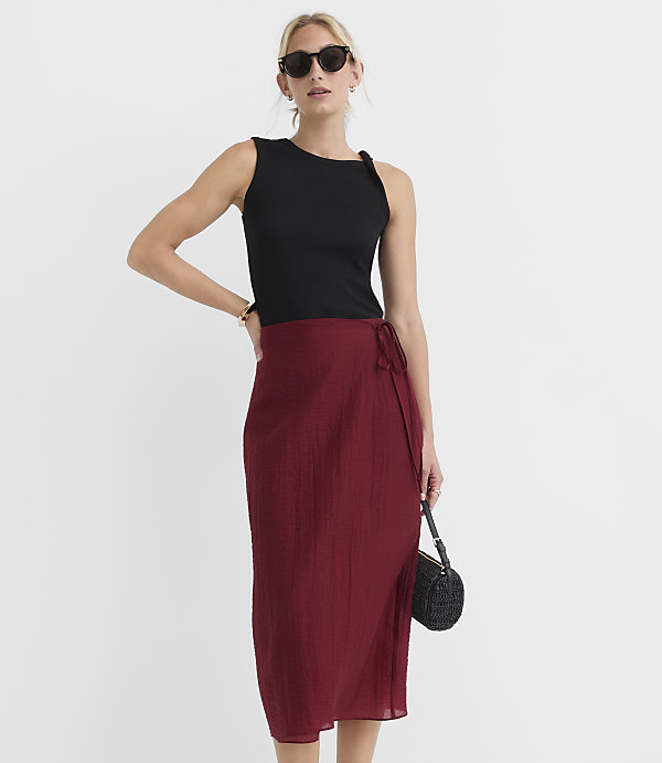A woman in a black tank top and a maroon wrap skirt poses against a plain white background, holding a small black beaded purse.