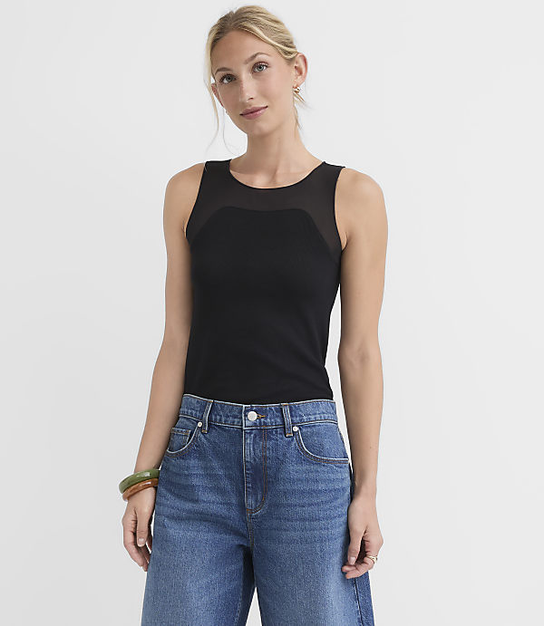 A woman in a black mesh-paneled tank top and blue jeans stands against a plain white background.