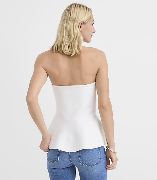 A woman in a white strapless peplum top and blue jeans stands against a plain white background.