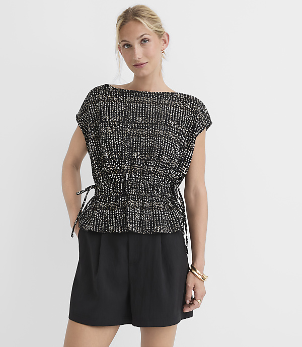 A woman in a patterned top and black shorts stands against a plain white background.