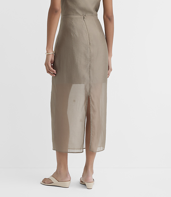 A woman in a sheer, taupe midi skirt and wedge sandals stands against a plain white background.