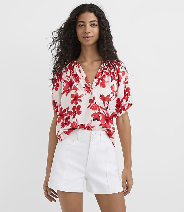 A woman in a floral blouse and white shorts stands against a plain white background.