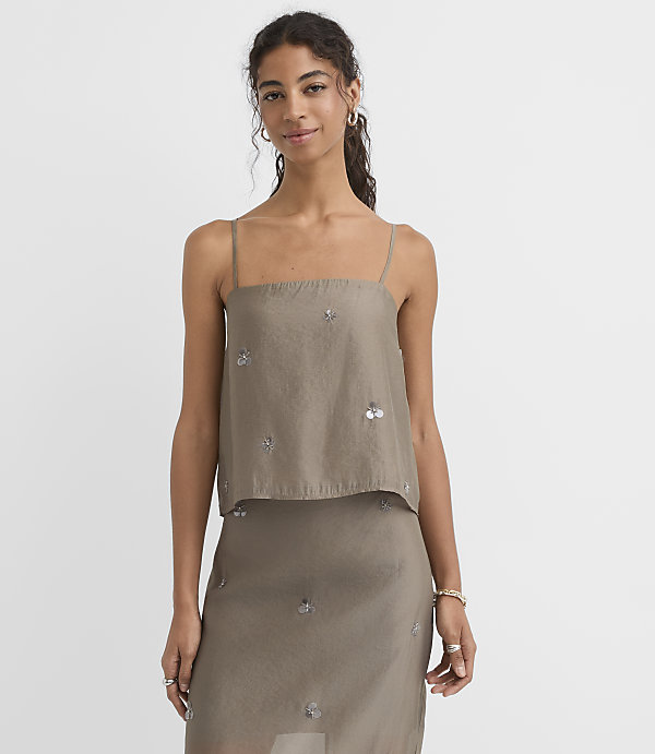 A woman in a taupe camisole top with sequin embellishments stands against a plain white background.