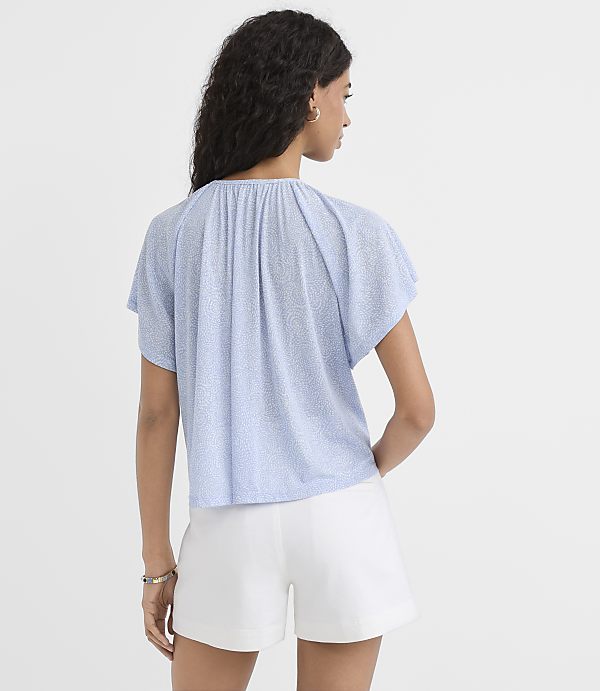 A woman in a light blue patterned top and white shorts stands against a plain light blue background.