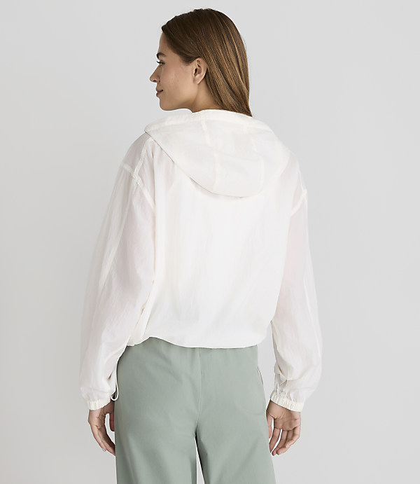 A woman in a white hooded jacket and green pants stands against a plain white background.