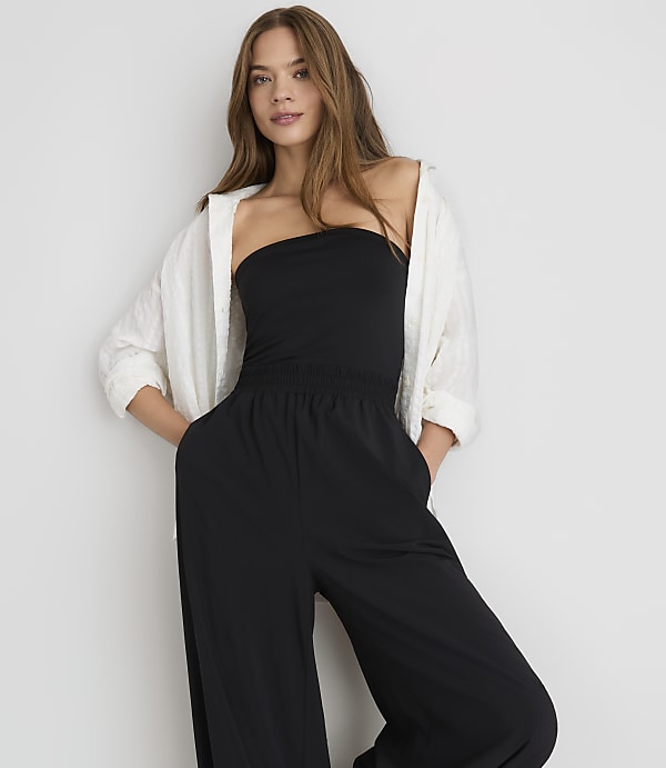 A woman in a black strapless jumpsuit and white open shirt stands against a plain white background.