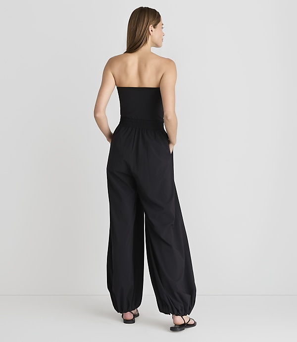 A woman in a black strapless jumpsuit stands with her hands in her pockets against a plain white background.