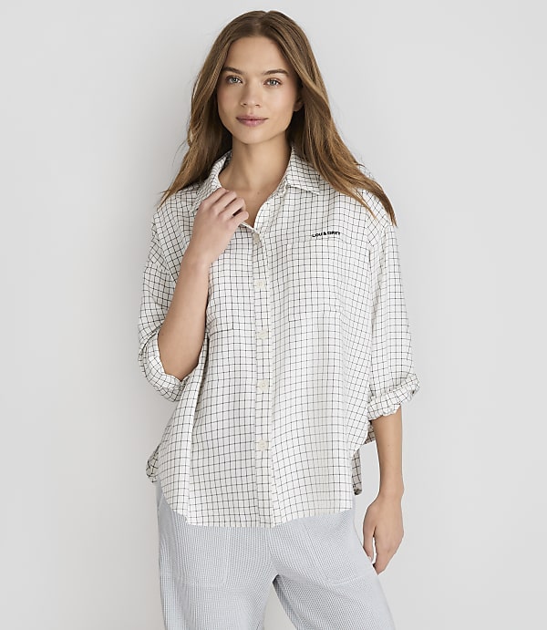 A young woman with long brown hair stands in the foreground wearing a checkered shirt and striped pants, against a plain white background.