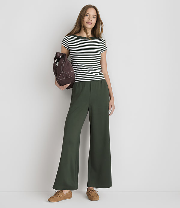 A woman in a striped shirt and wide-leg green pants stands against a plain white background, holding a brown bag.