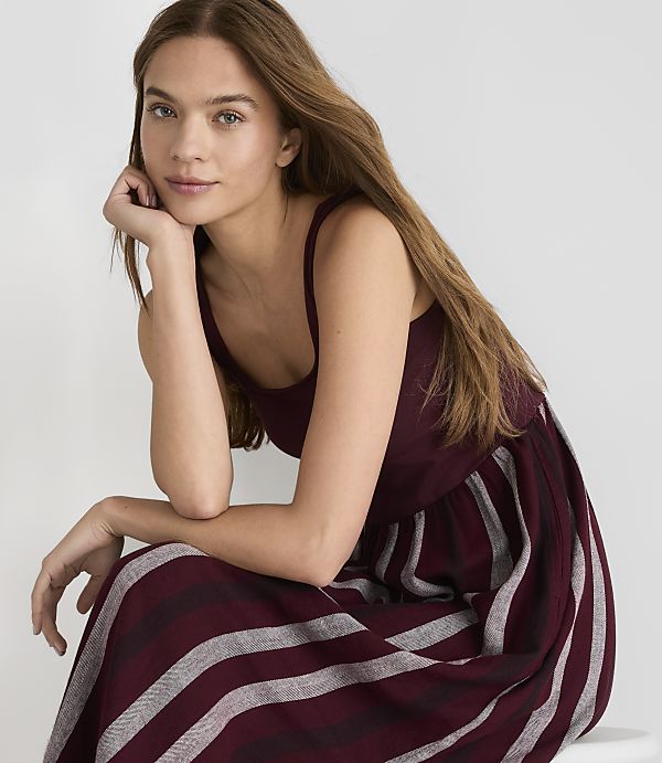 A young woman with long brown hair rests her chin on her hand, wearing a maroon and gray striped dress against a plain white background.