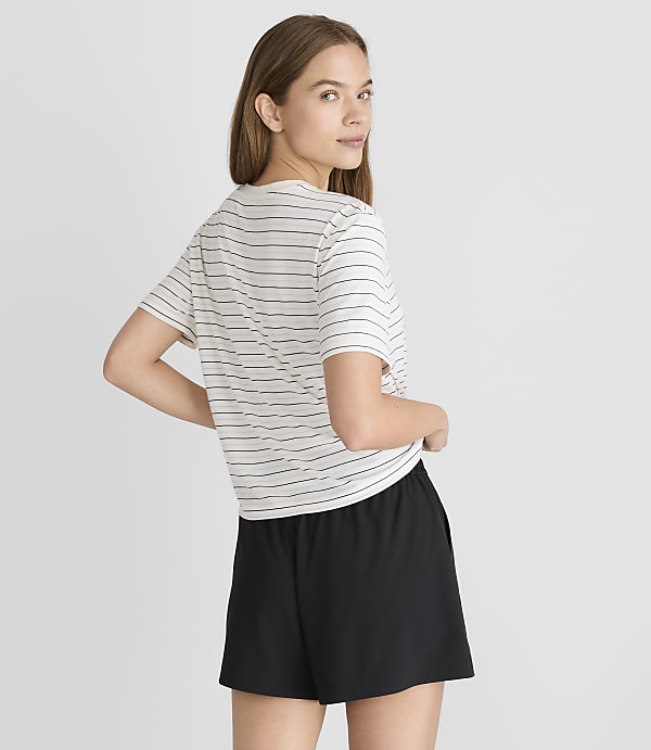 A young woman in a striped t-shirt and black shorts stands against a plain gray background.