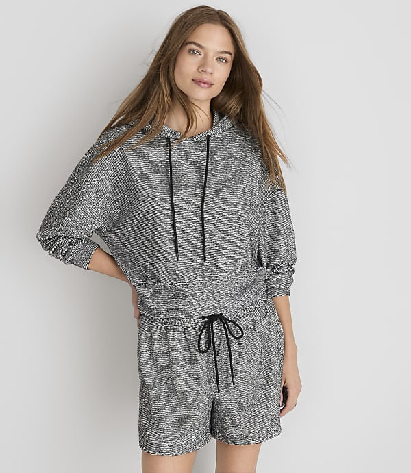 A young woman in a grey patterned hoodie and shorts stands against a plain white background.