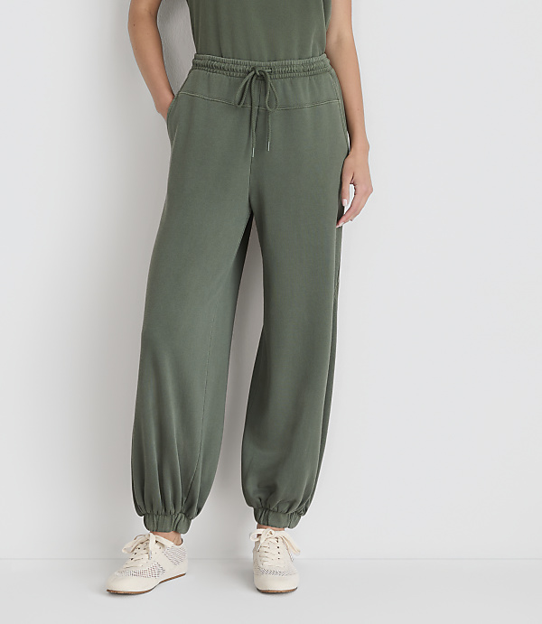 A person wearing olive green sweatpants and sneakers stands against a plain white background.